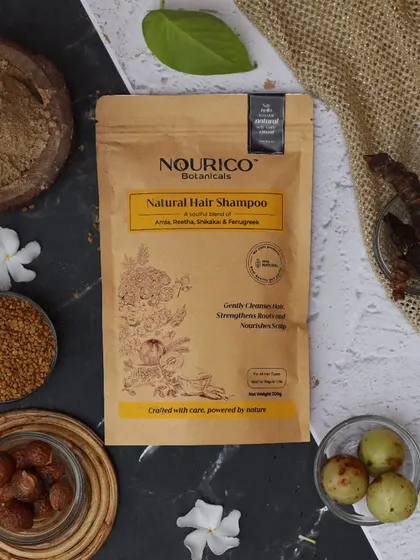 This flatlay tells the story of an Ayurvedic hair care brand through its raw ingredients. Amla, Reetha, and Shikakai are arranged around the product on a textured surface, emphasizing its natural and organic origins.