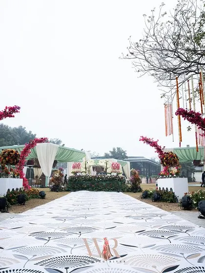 This mehndi entrance was a journey through a boho paradise. A custom-printed carpet leads the way, flanked by arches adorned with vibrant pink bougainvillea and consoles overflowing with colorful flowers, setting a joyful and spirited tone.