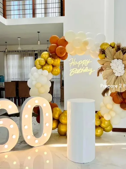 A warm and rustic-chic 60th birthday decoration. This setup features an arch of white, gold, and burnt orange balloons, a large dried palm leaf floral piece, a neon sign, and marquee numbers.