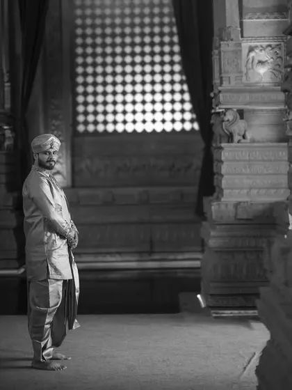 A groom stands in a sliver of light within a dark, cavernous temple hall. This dramatic use of shadow and light creates a moody, contemplative, and powerful black and white portrait.