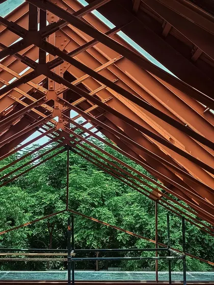 The intricate steel truss system of a roof is exposed during construction, revealing the geometric beauty of the building's skeleton against the green canopy.