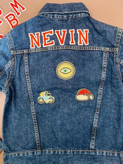 A custom denim jacket for 'Nevin', featuring his name in bold red letters. The jacket is decorated with unique patches, including an evil eye, a bicycle, and a Santa hat.