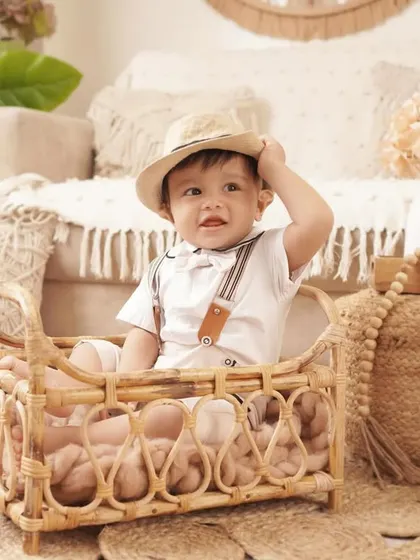 A playful shot of a baby boy tipping his hat while sitting in a wicker bassinet during his charming, boho-themed first birthday shoot.