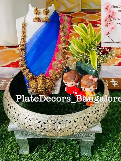A beautiful temple jewellery set displayed on a half-moon shaped platter. The arrangement includes cute couple dolls and fresh plants.
