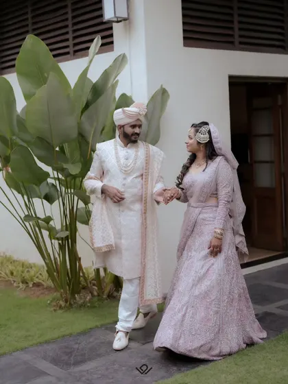 A couple walks hand-in-hand in a garden, dressed in elegant pastel wedding attire.