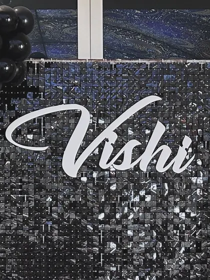 A personalized name cutout in a stylish script font, placed on a black shimmer wall. This is a great way to add a custom touch to a sophisticated party backdrop.