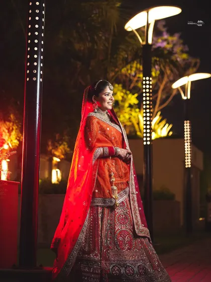 A smiling bride in her vibrant orange lehenga, posing outdoors at night. The modern lighting of the venue contrasts beautifully with her traditional attire.