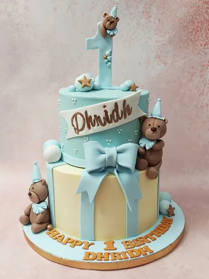 We design a classic and cuddly first birthday cake with this two-tiered teddy bear theme. Dressed in party hats and surrounded by blue and cream tones with a large bow, these teddy bears create a timeless and heartwarming centerpiece.