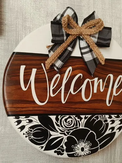 A classic "Welcome" sign to greet your guests. This piece combines a rich wood finish with bold floral graphics and a charming checkered bow, making any doorway feel more inviting.
