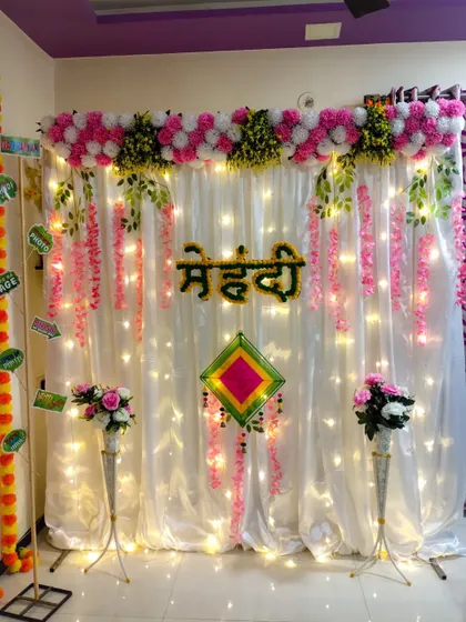 This simple and sober Mehndi function decor is perfect for intimate home events. It features a white backdrop with fairy lights, a pink and white floral header, and hanging pink flowers.