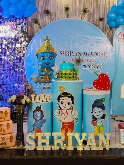 A grand Krishna-themed first birthday for Shriyan. The decor features a silver sequin wall, multiple character cutouts, and a blue and purple balloon arrangement.