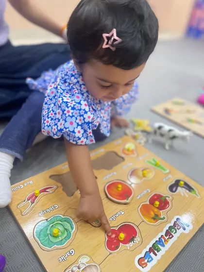 A toddler points to a tomato on a vegetable puzzle, learning the names of different foods and practicing the pincer grasp needed to handle the puzzle pieces.