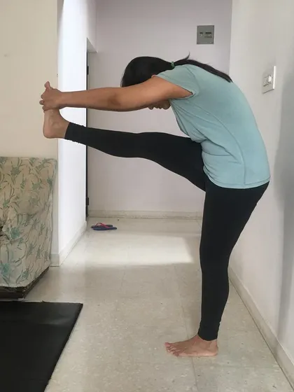 A student practices Dandayamana Janushirasana (Standing Head-to-Knee Pose) with wall support. In online classes, we use simple props available at home to make poses more accessible.