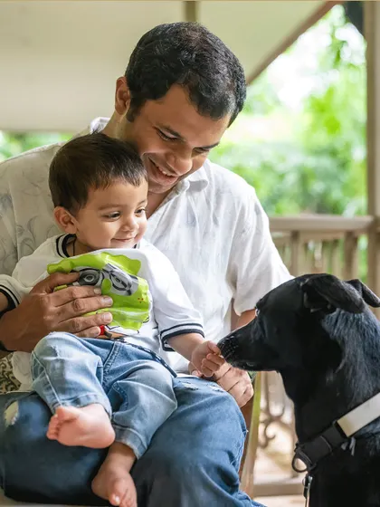 A father holds his smiling son on his lap while their black indie dog, Rio, comes in for a gentle sniff.