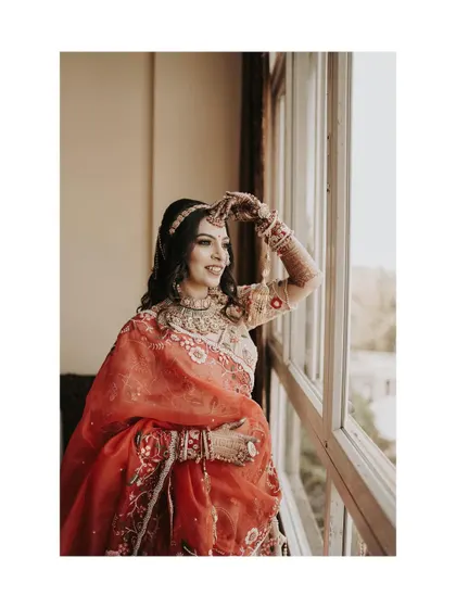 A classic bridal portrait by the window. The bride looks out, full of anticipation, her red and gold outfit glowing in the natural light.