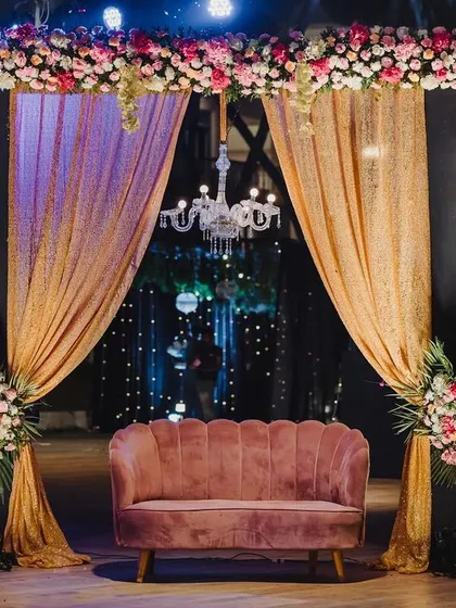 A cozy and elegant photo booth for a sangeet, featuring a plush pink sofa, a crystal chandelier, and shimmering golden drapes.