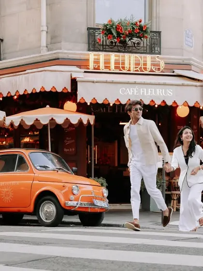 The couple joyfully crossing a street in front of a classic Parisian cafe and a vintage car, a picture-perfect moment of European romance.