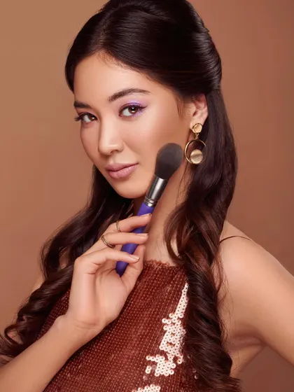 A glamorous campaign shot for a line of makeup brushes. The model holds a powder brush to her cheek, her pose exuding confidence and elegance. The warm tones and sparkling top create a festive, celebratory mood, perfect for a product line named "Dazzle."