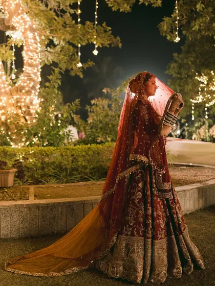 A classic bridal pose, showcasing the stunning details of her lehenga and the long, flowing veil. The garden setting at night adds a touch of romance and drama.