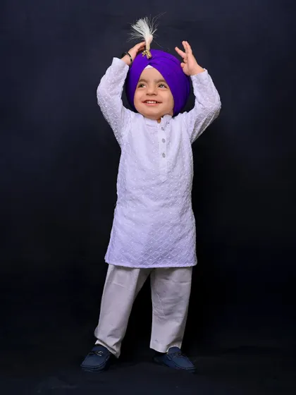 A young Sikh boy in a white kurta and a vibrant purple turban smiles joyfully. This individual portrait, part of a family session, captures his bright and happy personality.