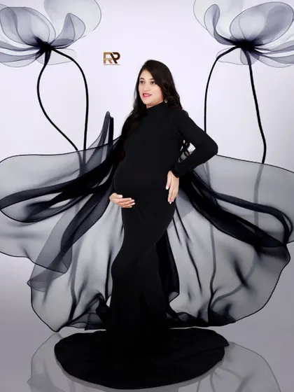 A high-fashion maternity portrait. The mom-to-be wears a sleek black gown, with sheer fabric creating a dramatic, wing-like effect. The giant floral props add an artistic, modern touch.