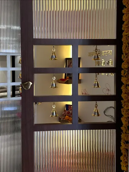 A close-up of a custom pooja room door. The design features frosted glass panels and small cutouts with traditional brass bells, creating a beautiful and spiritual entrance.