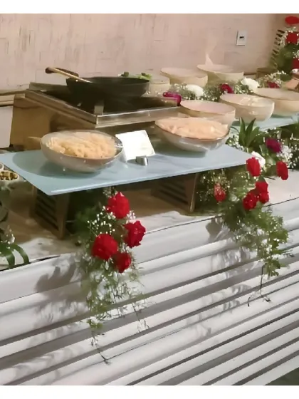 A live noodle station setup with ingredients neatly arranged in bowls on a glass-tiered stand. The station is decorated with cascading red flowers, adding a pop of color.