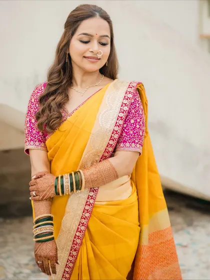 A full-length view of a simple yet stunning Marathi bridal look. The bride's yellow saree with a contrasting pink blouse is complemented by minimal makeup, soft waves, and traditional green bangles.