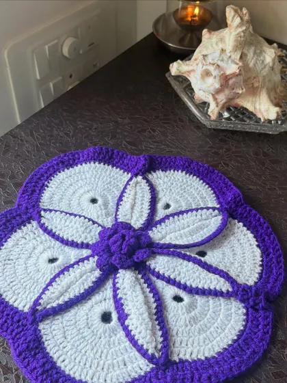 A beautiful crochet table mat with a purple and white floral petal design, adding an elegant and handmade accent to a side table.