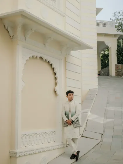 A solo portrait of the groom looking sharp in his traditional attire against the pristine white architecture of a Udaipur palace. It's a moment of quiet confidence before the big day.