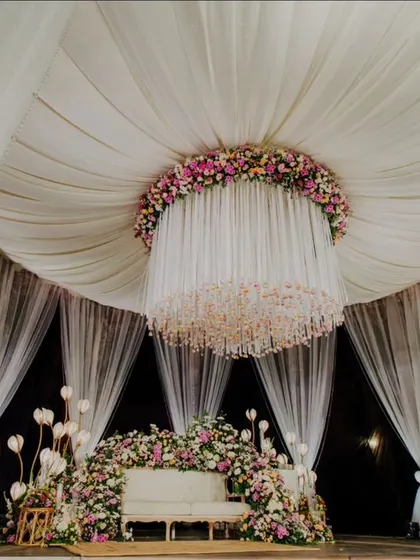 The reception stage is a vision in white, with dramatic draping, a stunning central chandelier, and a couch nestled in a garden of soft pink and white flowers.