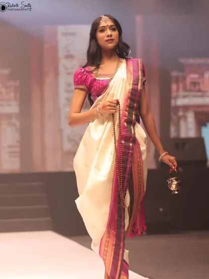 A model in a white and pink traditional outfit walks the runway. This full-length shot showcases the unique draping style and the simple elegance of the design.