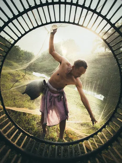 A fisherman casts his net, captured through the circular frame of a fishing basket. This is a masterful example of creative composition and lighting in street and documentary photography.