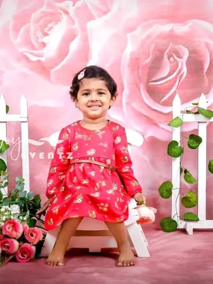 A lovely rose-themed kids' photoshoot. The little girl sits in front of a pink floral backdrop with a white picket fence, creating a sweet and classic garden party look.