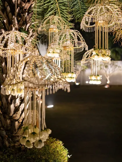 A magical detail from an outdoor event: intricate, kalire-style chandeliers hanging from a palm tree, illuminated for the evening.