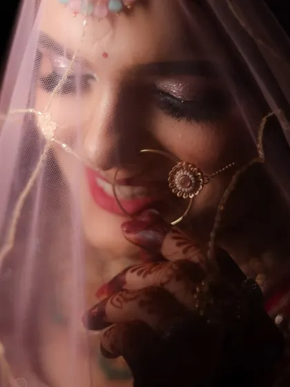 A soft, romantic shot of a bride peeking through her pink dupatta. The lighting highlights her smile and the subtle glitter on her eyes.