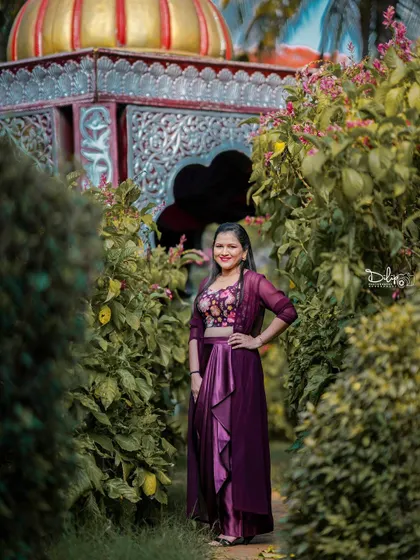 A solo portrait of a model in a beautiful purple dress, posing in a garden pathway with a gazebo in the background.