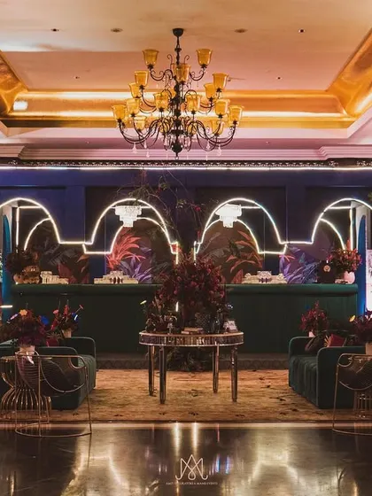 A full view of the contemporary sangeet hall, with a stunning bar, chic lounge seating, and a grand chandelier, all tied together with a bold color palette.