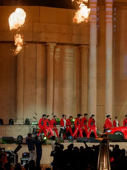 Pillars of fire erupt on stage during a performance, adding to the drama and spectacle of the event. The monumental columns of the stage provide a powerful, ancient-feeling backdrop.