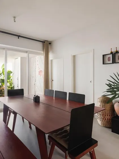 The dining area in the 'Rustic Minimal' apartment, featuring a large mahogany table and bench designed to host big family gatherings. The space opens directly onto a balcony, extending the living area and bringing in fresh air.