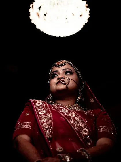 A dramatic low-angle shot of a bride in her wedding attire. The single light source from above creates a spotlight effect, focusing all attention on her serene expression and the rich details of her red lehenga.