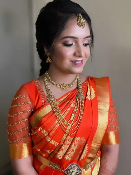 A lovely bride ready for her ceremony. Her hair is styled in a simple, elegant braid that complements her bright orange and gold silk saree and traditional jewelry.