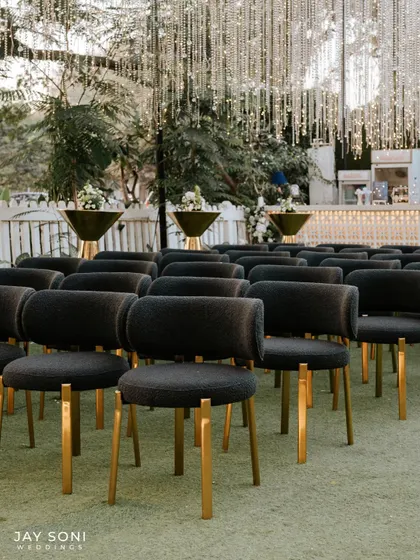 Modern and sophisticated seating for an engagement party. The dark, textured chairs with gold legs are arranged neatly, facing a bar area adorned with cascading string lights.