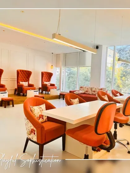 This shot shows the overall layout of the nail studio, from the pedicure stations with their comfortable high-back chairs to the manicure tables. The color palette of vibrant orange and clean white creates a feeling of playful sophistication. Large windows allow natural light to fill the space, making it feel bright and airy.