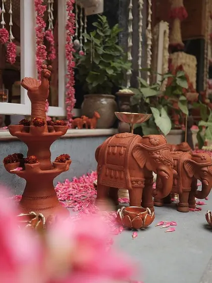 A low angle shot of terracotta elephant figurines and a traditional lamp, with pink flower petals scattered on the floor. This composition highlights the rich textures and cultural significance of the decor.
