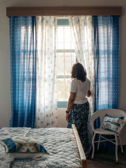 A moment of reflection by the window. The layered blue curtains, one with a geometric print and the other with a delicate buti pattern, create a stunning visual frame and a peaceful mood.