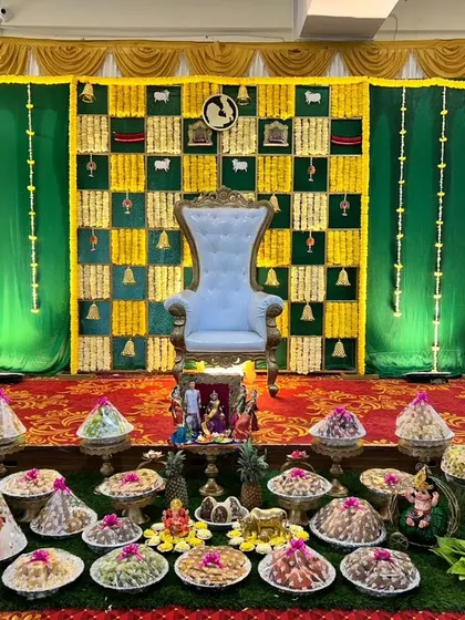 A traditional green and yellow themed Seemantham decor. The stage includes a floral backdrop, a ceremonial throne, and a wide array of beautifully arranged thamboolam plates.