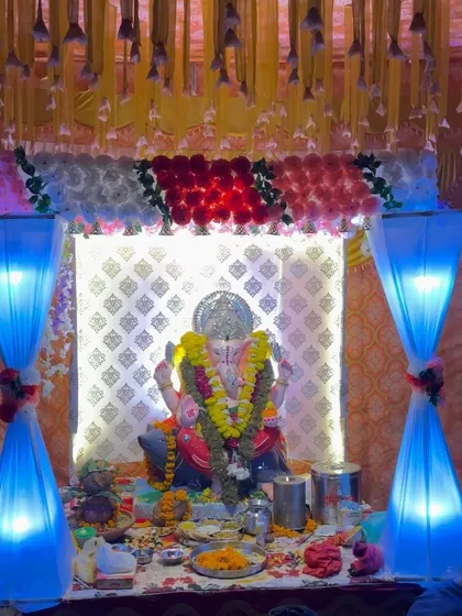 A Ganpati puja setup with blue-lit fabric columns. The idol is placed against a decorative white backdrop, with a floral border on top.