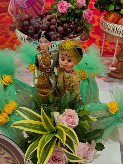 An adorable couple doll arrangement perfect for an engagement or wedding. The dolls are dressed in traditional attire and placed amidst fresh flowers and a platter of grapes.