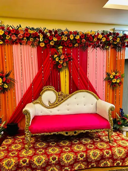 This is a classic wedding stage I set up for a reception in Gurgaon. It features a backdrop of bright red, orange, and pink drapes, a thick garland of fresh gerberas and other flowers, and a traditional white and gold sofa for the couple.